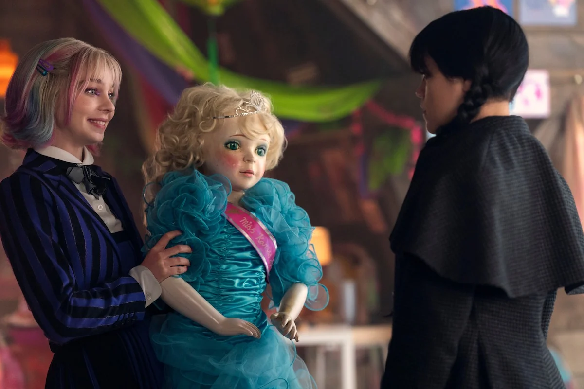 Emma Myers as Enid Sinclair holds a doll and smiles at Jenna Ortega as Wednesday Addams in Wednesday Season 2